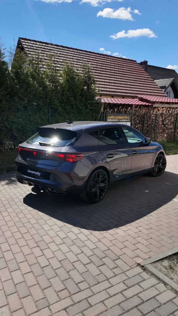 Szary hatchback marki Cupra zaparkowany na utwardzonym podjeździe przed domem.