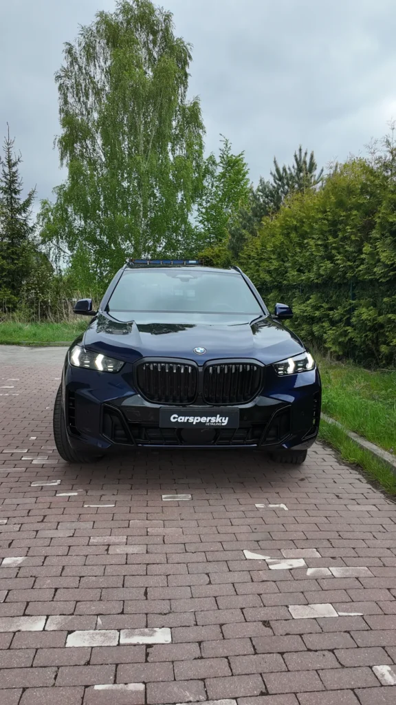 Ciemnoniebieski SUV marki BMW zaparkowany na utwardzonym podjeździe, z logo Kaspersky na przednim zderzaku.