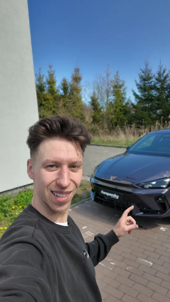 Mężczyzna w czarnej bluzie uśmiecha się i wskazuje na ciemny samochód marki Cupra zaparkowany na utwardzonym parkingu, podkreślając jakość jego folii PPF.